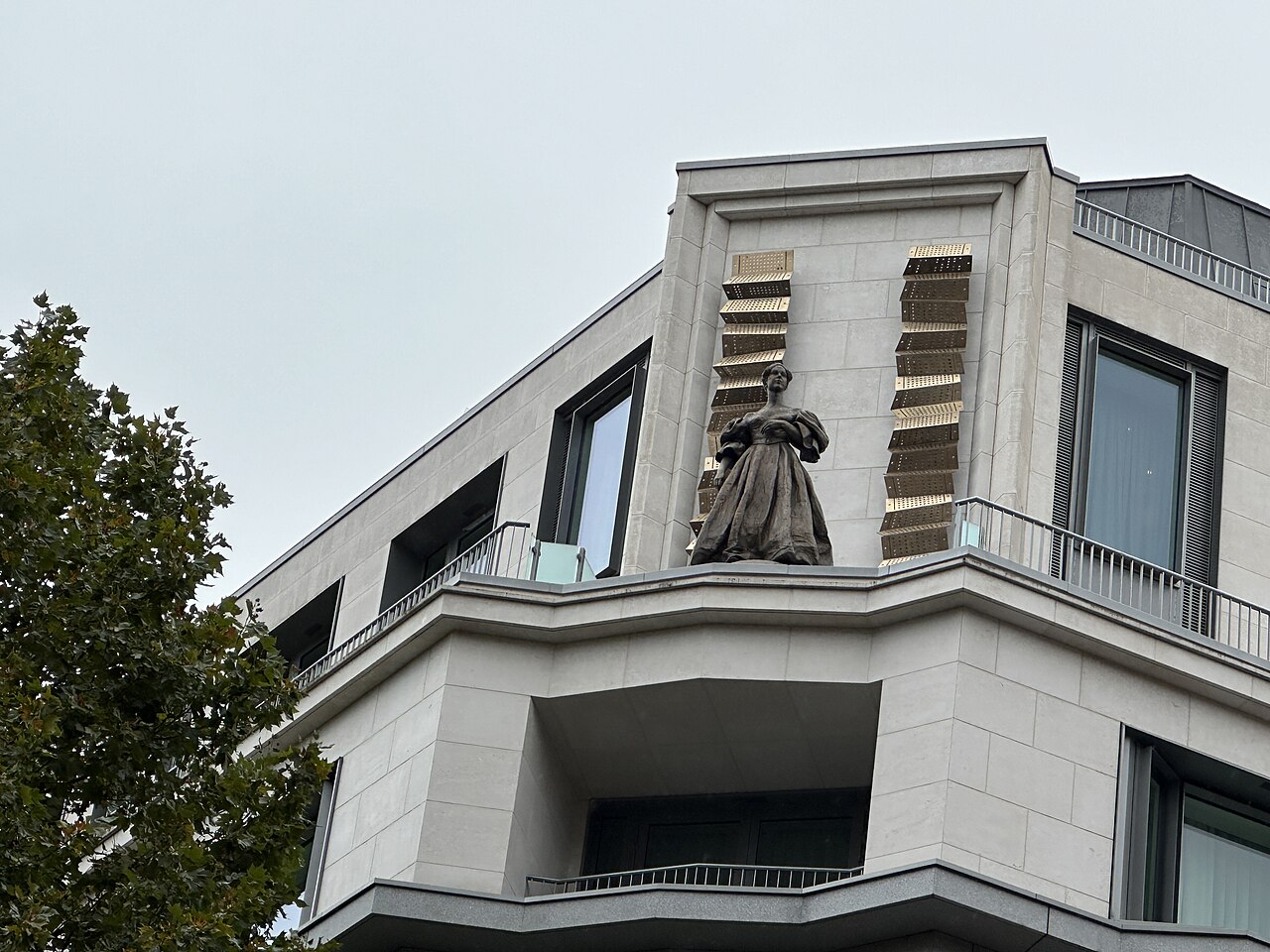 Estátua de bronze de Ada Lovelace de vestido longo, no alto de um edifício claro, entre duas colunas douradas de cartões perfurados dobrados.