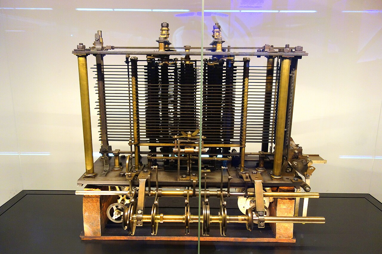 Peça de teste da Máquina Analítica: estrutura de latão com fileiras de hastes horizontais e eixos de engrenagens, sobre uma base escura, dentro de uma vitrine.