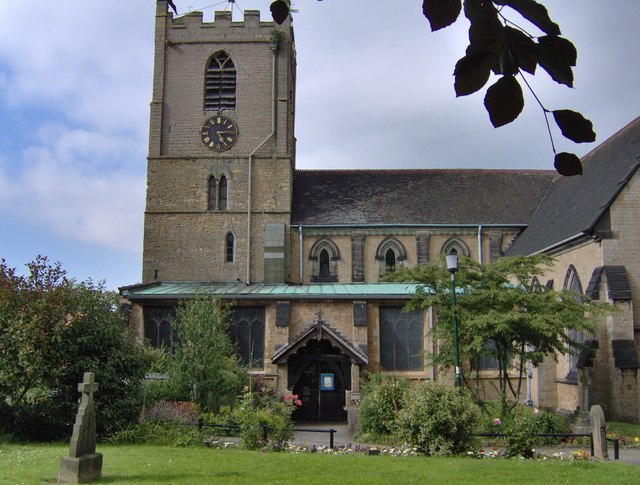 Igreja de pedra de St Mary Magdalene, em Hucknall, com torre de relógio, entrada com pórtico, gramado e uma cruz de pedra em primeiro plano.