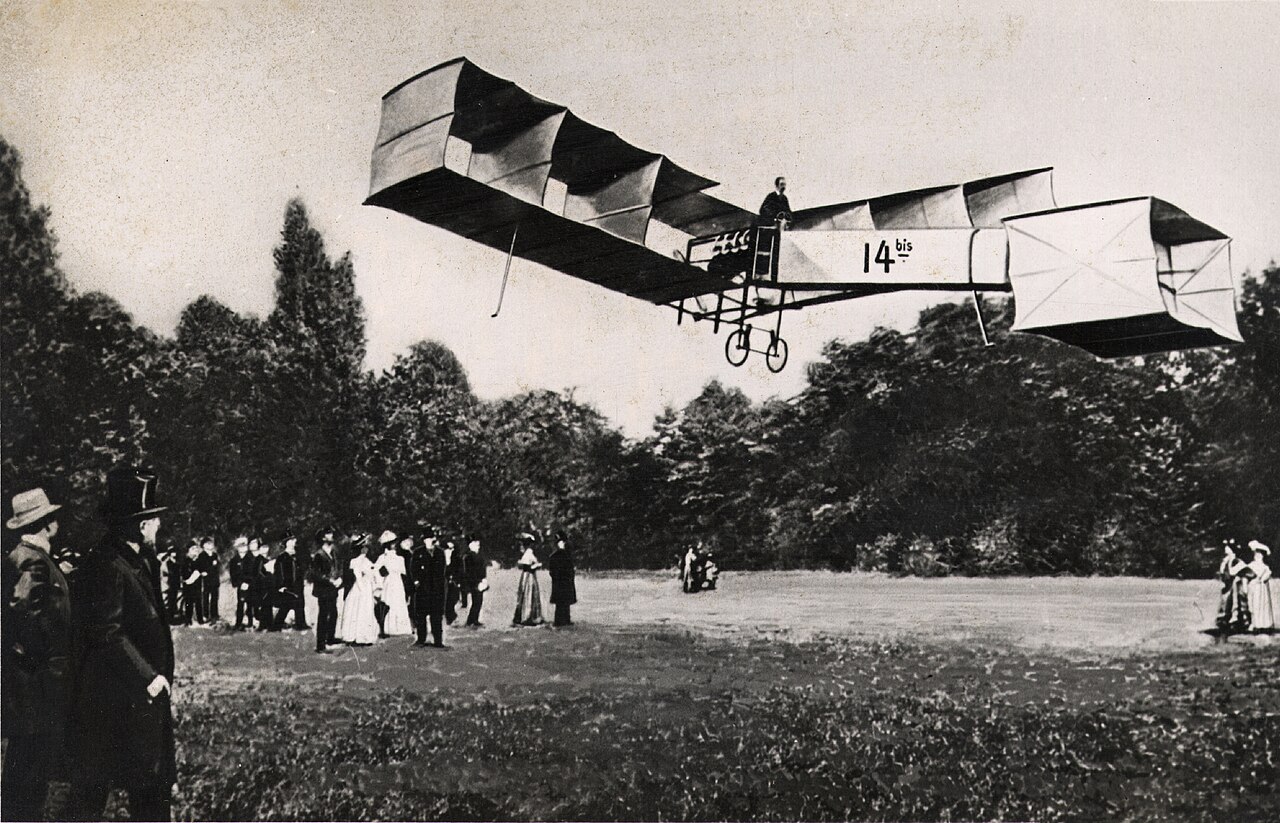 O 14-bis, aeroplano de caixas de asas com o número 14 bis na lateral, no ar sobre o gramado de Bagatelle, com um público ao fundo.