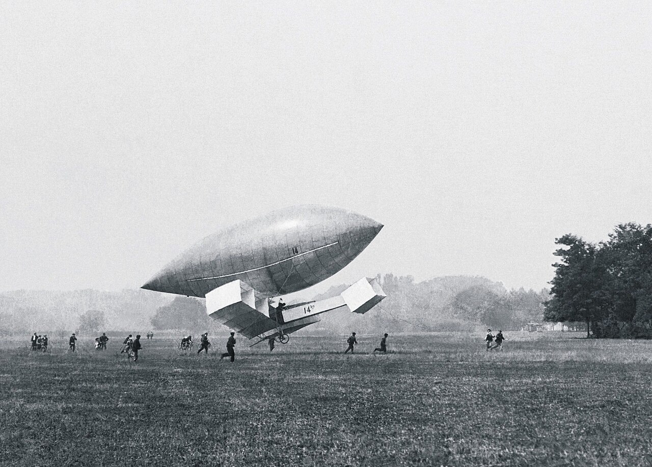 Fotografia antiga de um aeroplano de caixas de asas pendurado sob um dirigível alongado, sobre um campo plano, com pessoas correndo embaixo.