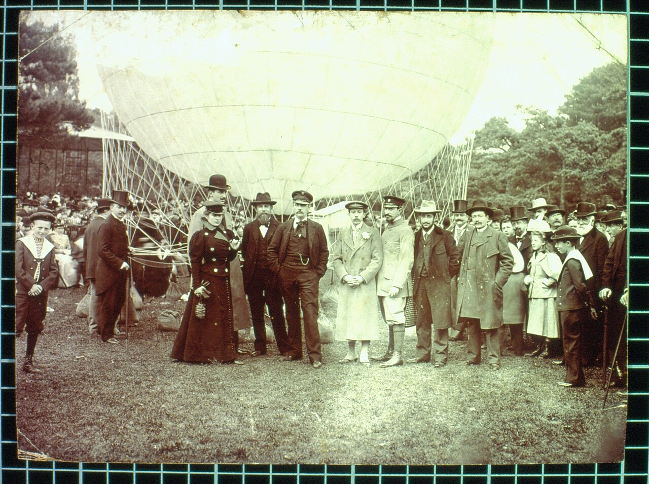 Fotografia antiga de um grupo de homens e mulheres em pé diante de um grande balão, com a cesta ao lado, num parque.