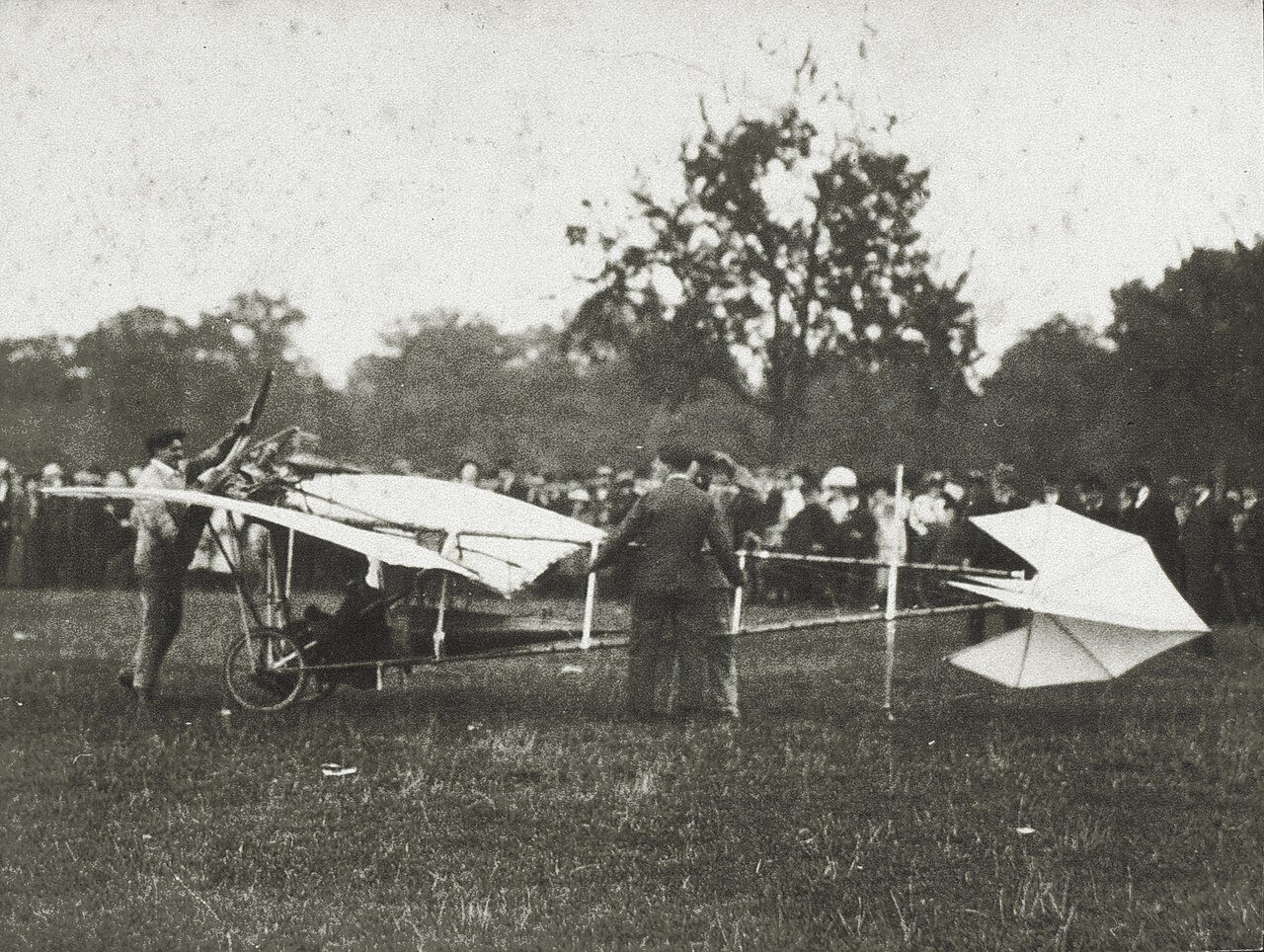 Fotografia antiga da Demoiselle, monoplano leve de asas longas e cauda em losango, parado num campo com muitas pessoas ao fundo.