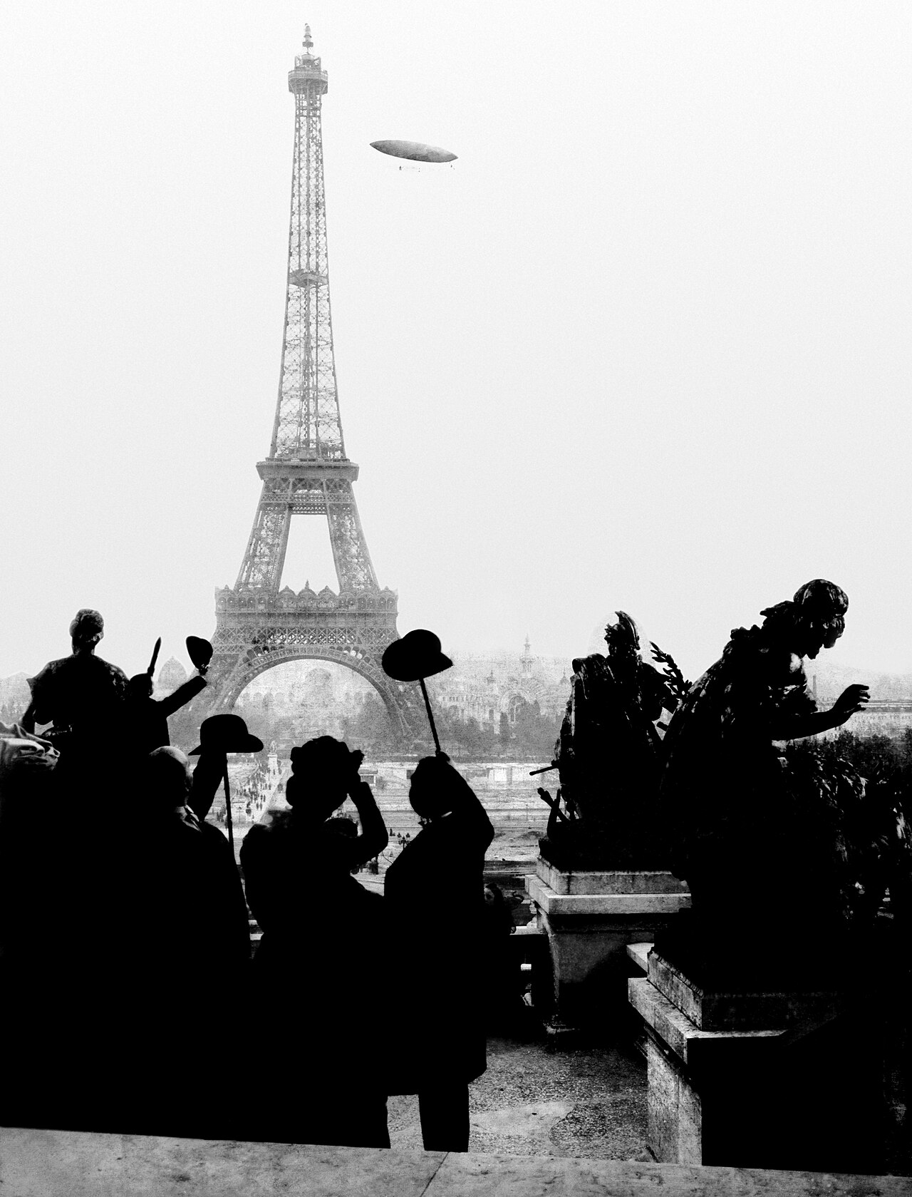 Silhueta de pessoas acenando em primeiro plano, a Torre Eiffel ao centro e, ao fundo, no céu, o dirigível de Santos-Dumont.