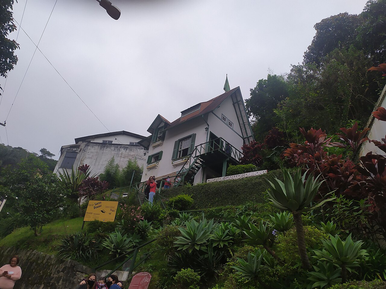 Casa de dois andares com varanda no alto de um morro verde, entre palmeiras e plantas, vista de baixo, da rua, em Petrópolis.