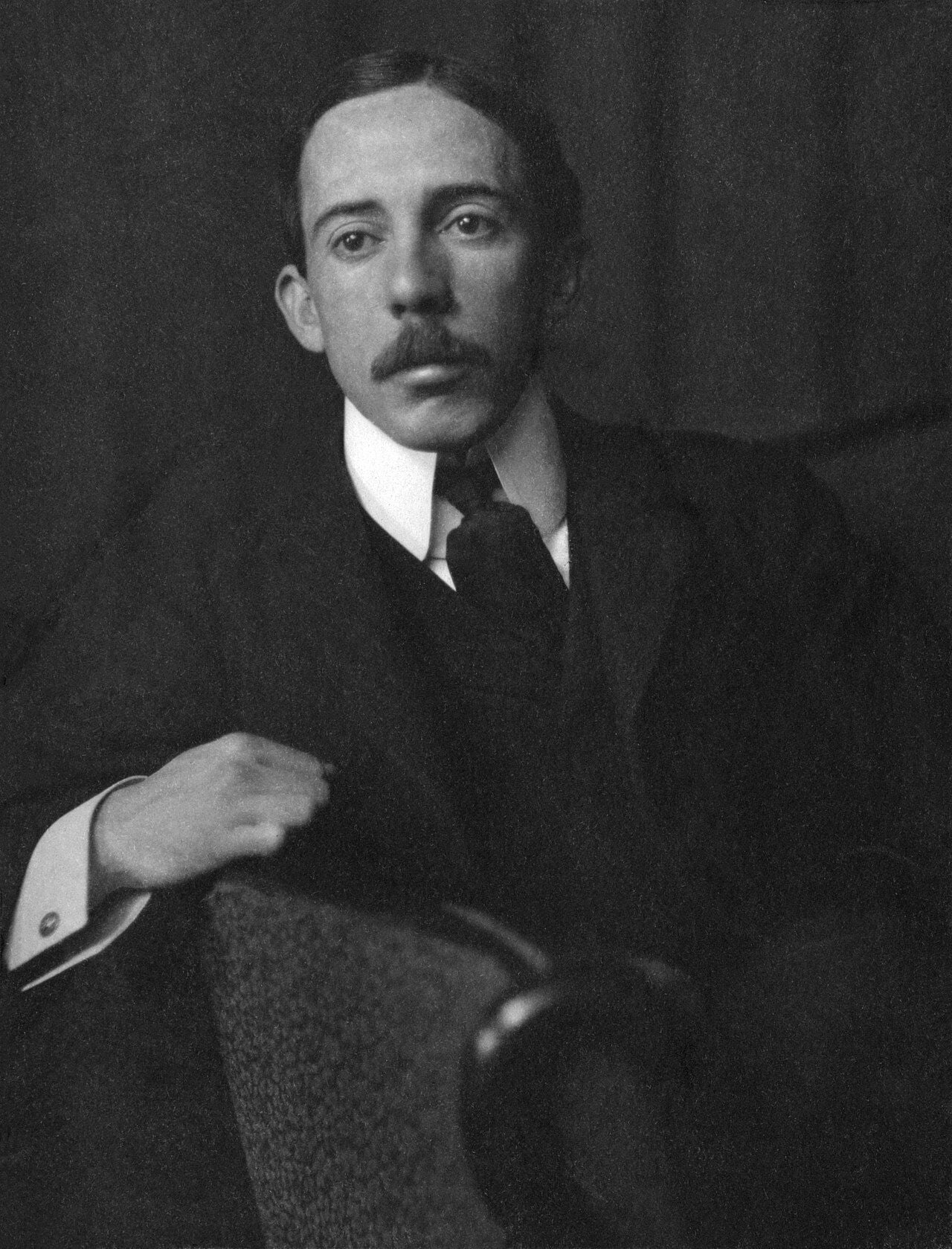 Retrato em preto e branco de Alberto Santos-Dumont, sentado, de terno e gravata, com o braço direito apoiado no encosto da cadeira.
