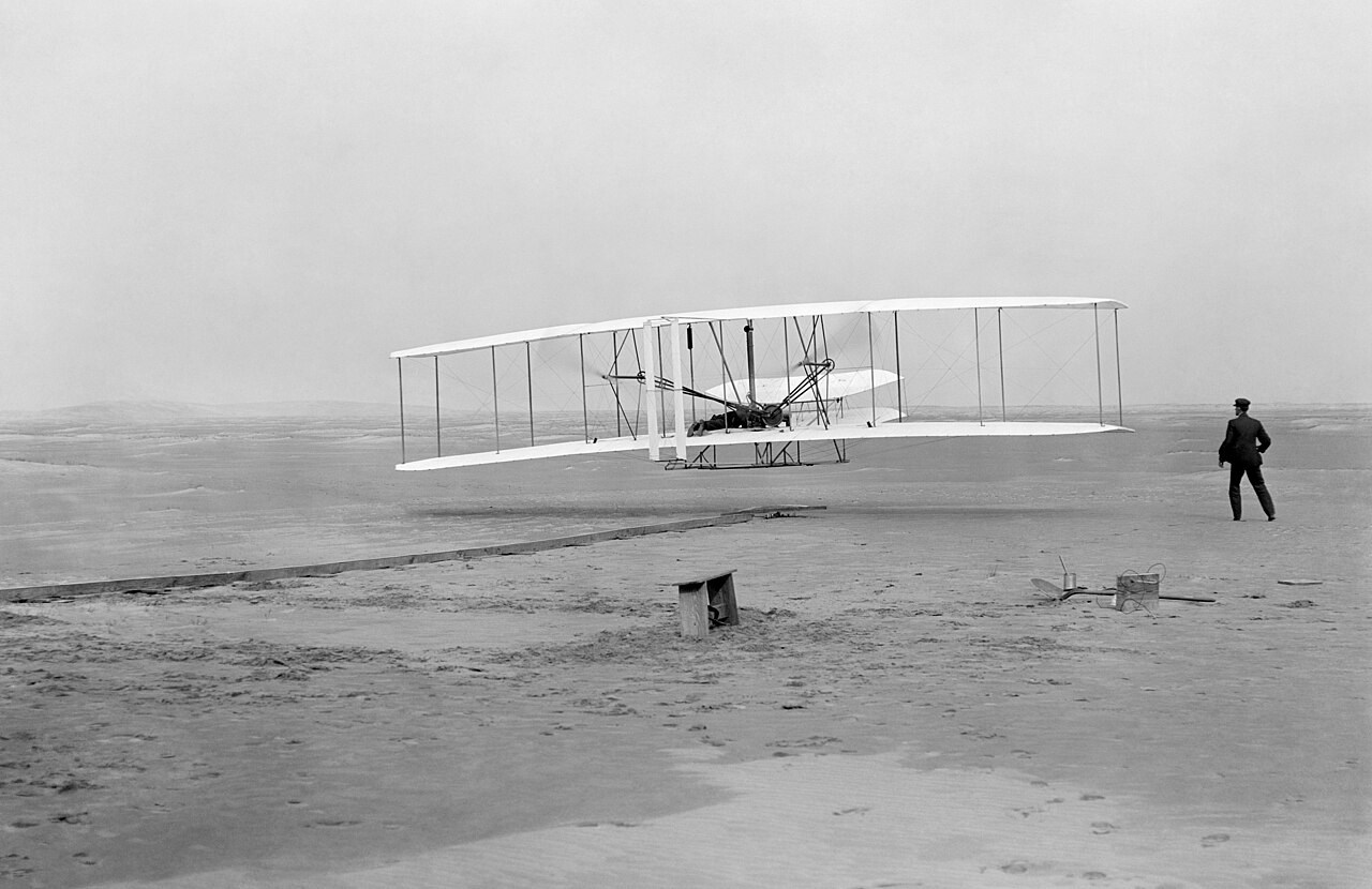 Foto em preto e branco do Flyer dos irmãos Wright poucos metros acima da areia de Kitty Hawk, com um homem em pé ao lado, à direita.