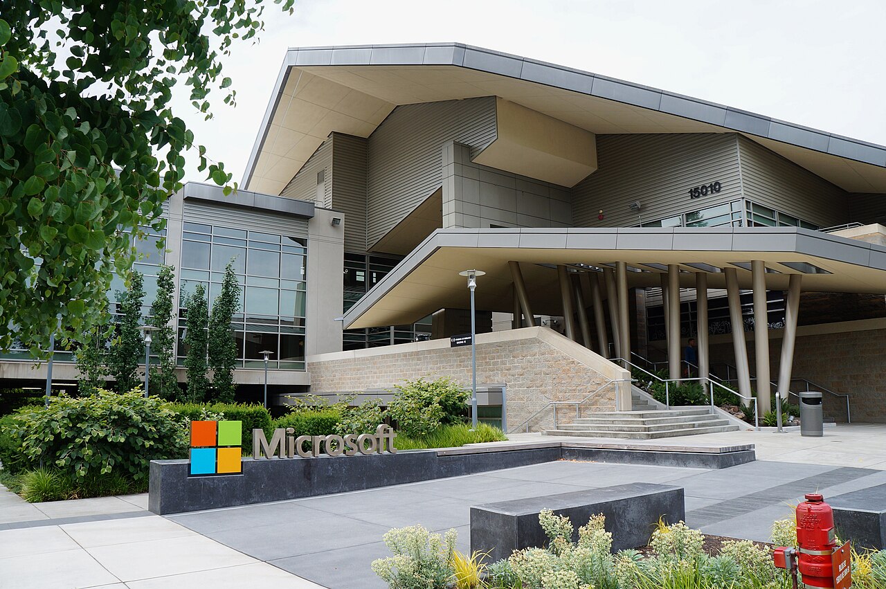 Entrada de um edifício moderno de telhado inclinado sobre colunas, com o letreiro Microsoft num canteiro à frente.