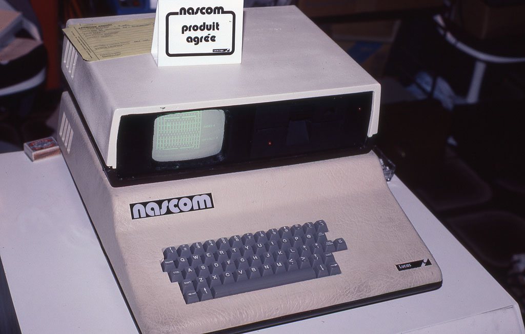 Computador Nascom 2 bege, com teclado cinza, monitor pequeno e unidade de disquete, sobre uma mesa branca, com um cartaz da marca acima.