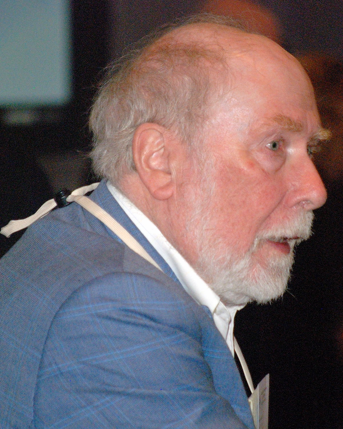 Niklaus Wirth, de perfil, com cabelo e barba brancos, paletó azul e um cordão no pescoço, falando num evento.