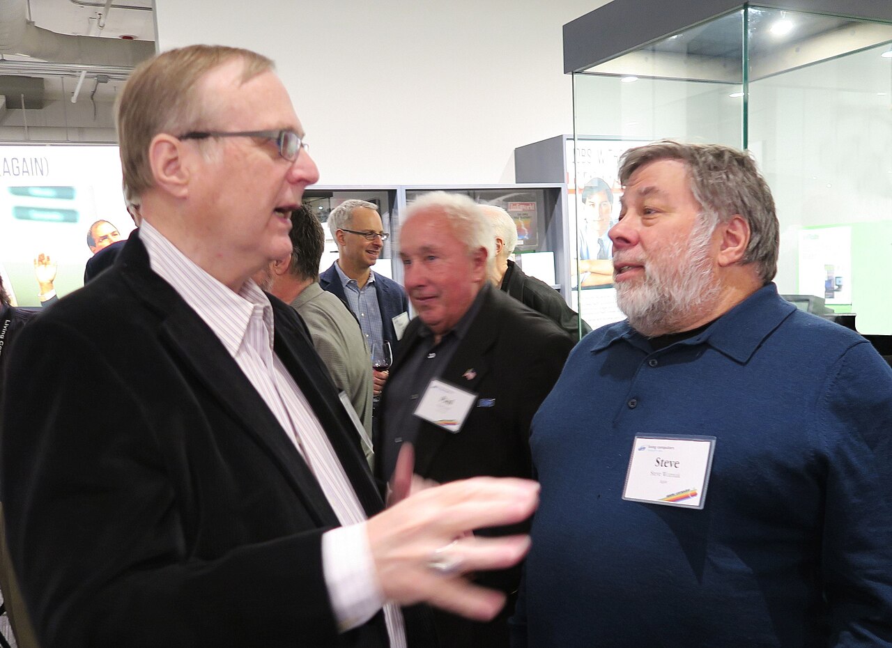 Paul Allen, de paletó escuro e óculos, conversando com Steve Wozniak, de barba grisalha e camisa azul-marinho com crachá, num museu com vitrines de vidro.