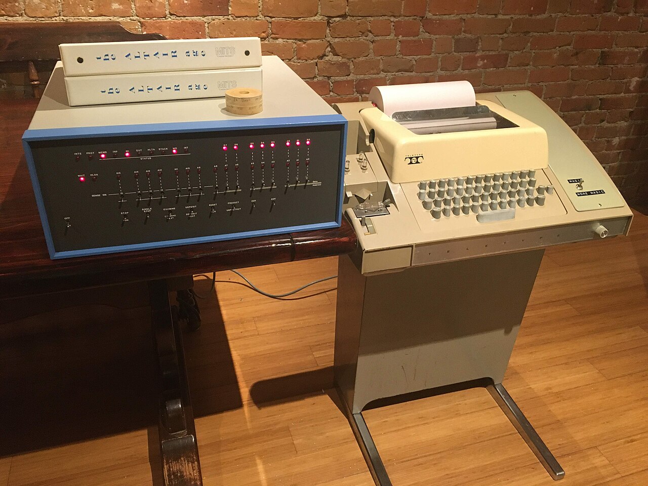 Computador Altair 8800 de painel azul e cinza, com interruptores e luzes vermelhas, ao lado de um terminal Teletype bege sobre pedestal, com um rolo de fita de papel sobre o computador.