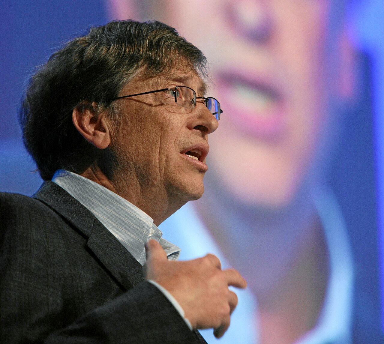 Bill Gates de perfil, de óculos e paletó escuro, falando e gesticulando com a mão no peito, diante de uma tela com o rosto dele ampliado e desfocado.