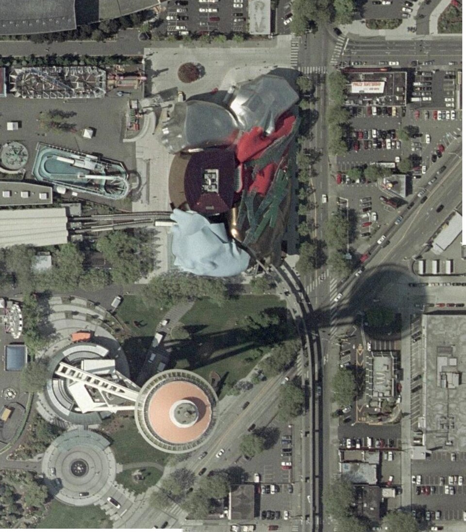 Vista aérea do Seattle Center: o prédio de formas curvas e metal colorido do Experience Music Project ao lado da Space Needle, cuja sombra se estende sobre ruas e estacionamentos.