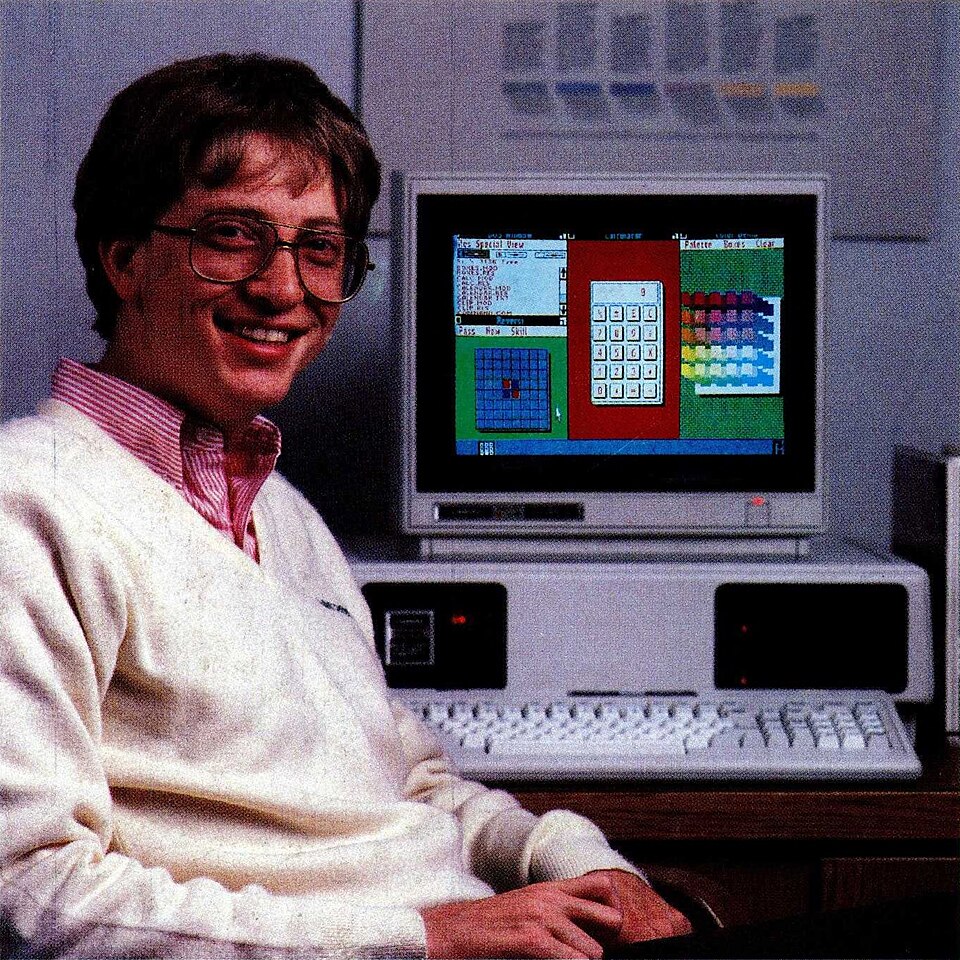 Bill Gates jovem, de óculos e suéter claro, sorrindo para a câmera, sentado diante de um computador de mesa cuja tela mostra janelas lado a lado, com uma calculadora ao centro.