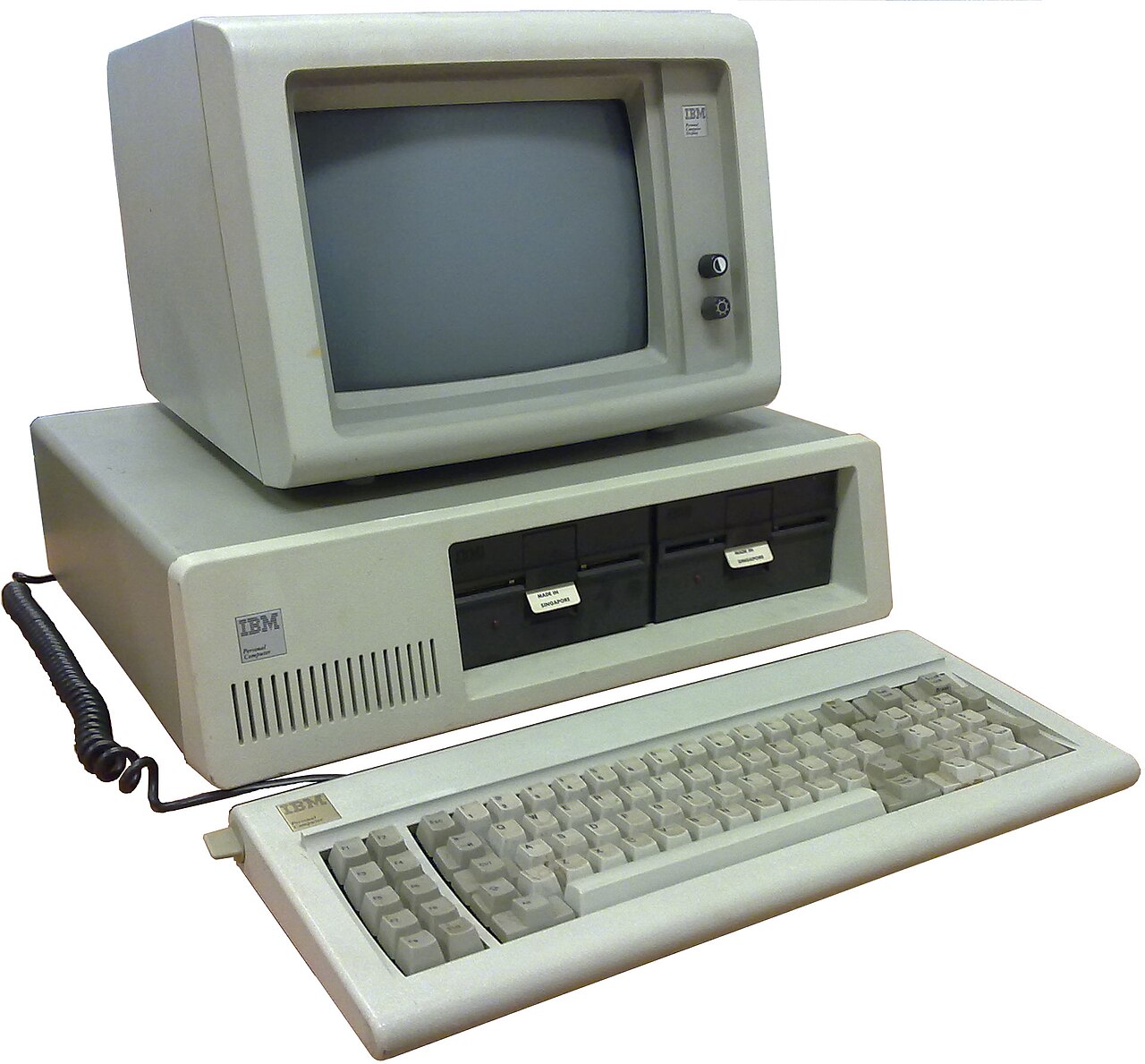 Computador IBM PC modelo 5150: caixa bege com duas unidades de disquete, monitor monocromático em cima e teclado à frente, ligado por um cabo espiralado.