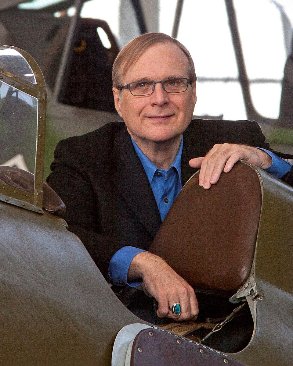 Paul Allen de paletó escuro e camisa azul, sorrindo levemente, apoiado no assento de um avião antigo de cabine aberta, em um hangar.