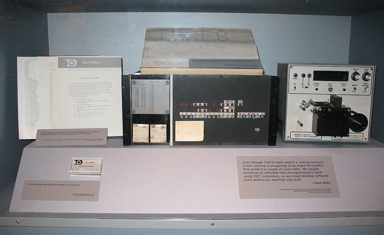 Vitrine de museu com o computador Traf-O-Data, uma caixa preta com fileiras de luzes vermelhas, ao lado de um leitor de fita de papel, folhetos, um cartão de visita e placas com texto.