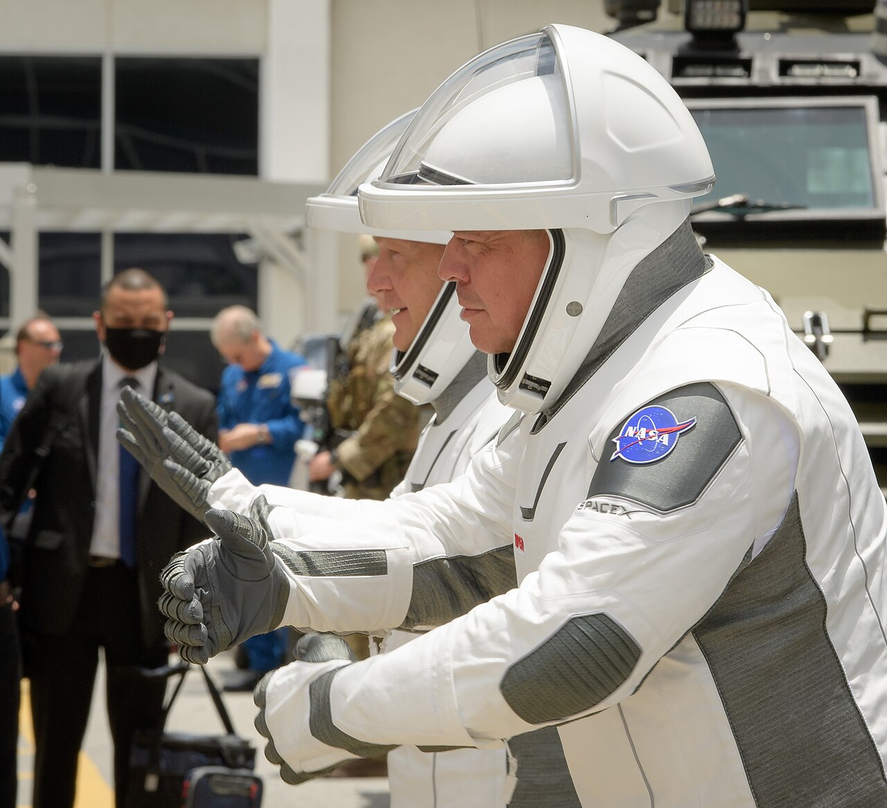 Dois astronautas de traje espacial branco e cinza, com capacete, fazem gestos com as mãos diante de pessoas que aparecem ao fundo.