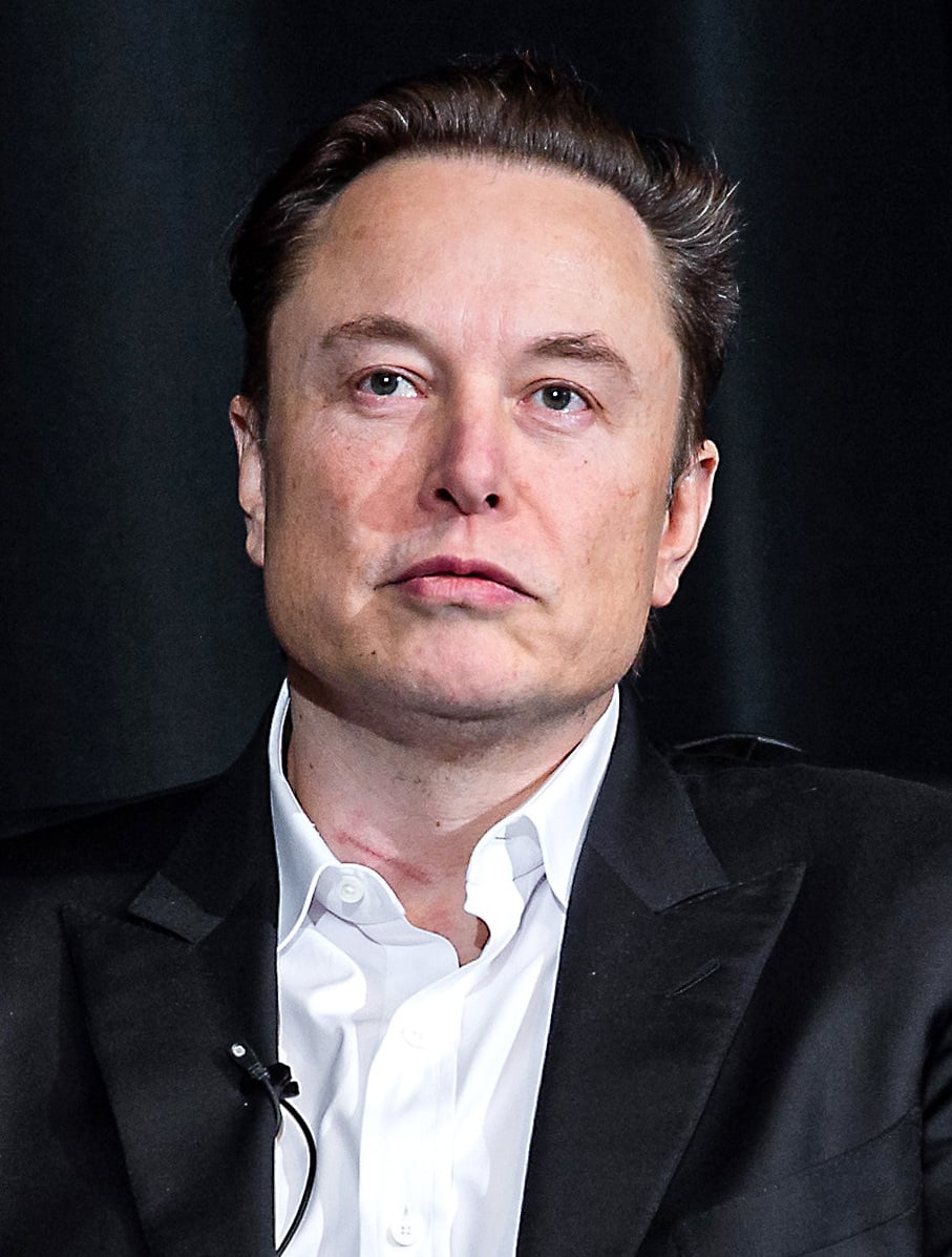 Retrato de Elon Musk, de terno escuro e camisa branca aberta no colarinho, com um microfone de lapela, diante de um fundo escuro.