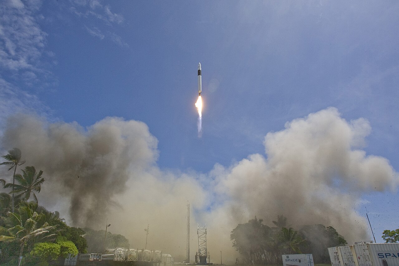 Um foguete Falcon 1 sobe com uma chama brilhante, entre nuvens de fumaça e poeira e palmeiras, num céu azul.