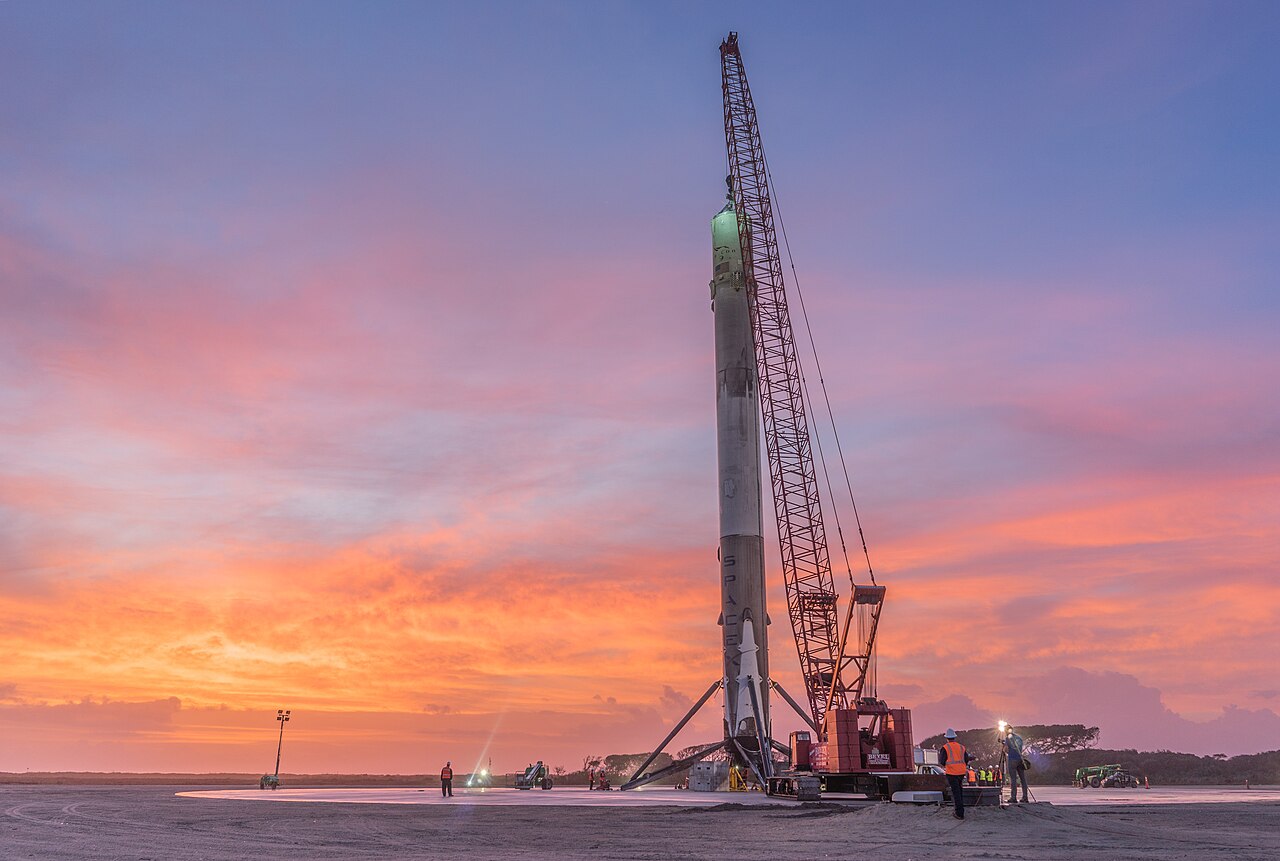 Primeiro estágio de um foguete Falcon 9 em pé sobre suas pernas no solo, ao lado de um guindaste, ao amanhecer, com céu rosa e laranja.