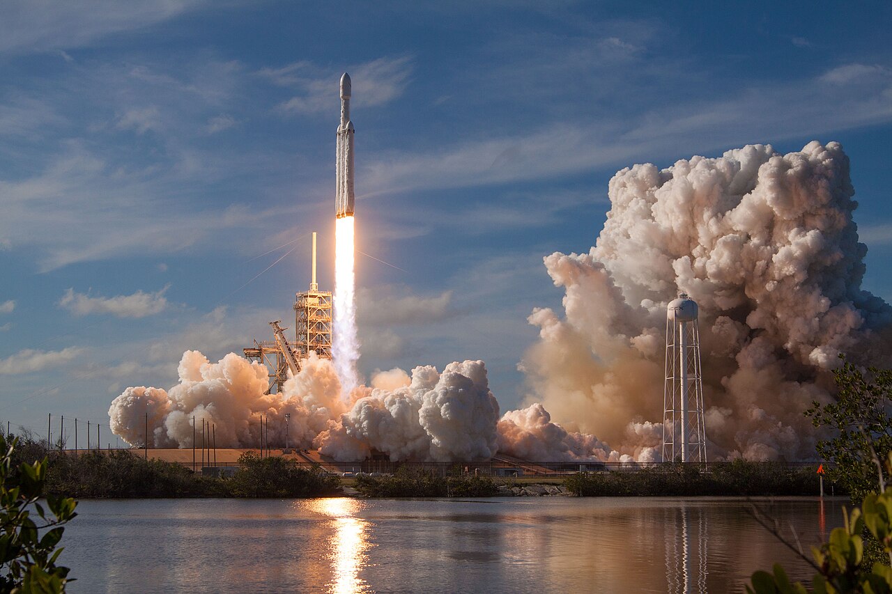 O foguete Falcon Heavy subindo com uma longa chama ao lado de uma torre de serviço, entre grandes nuvens de vapor, com um lago em primeiro plano.