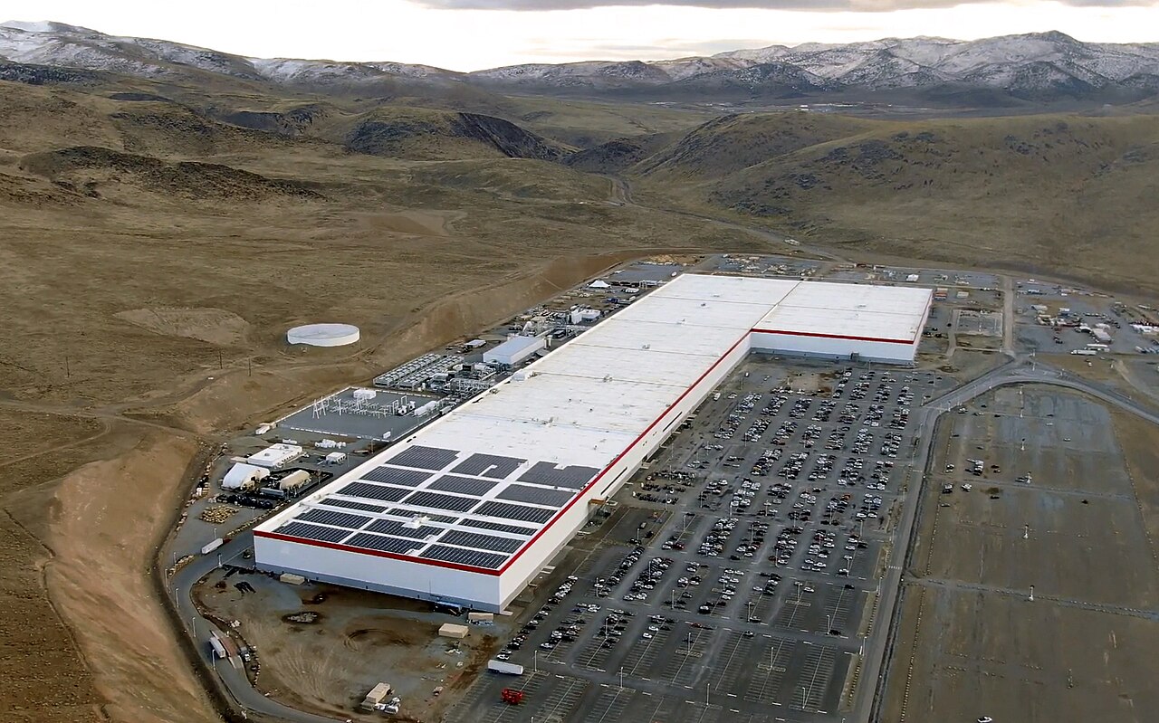 Vista aérea de um prédio industrial branco, enorme e comprido, com painéis solares no telhado, cercado por estacionamento e colinas áridas.