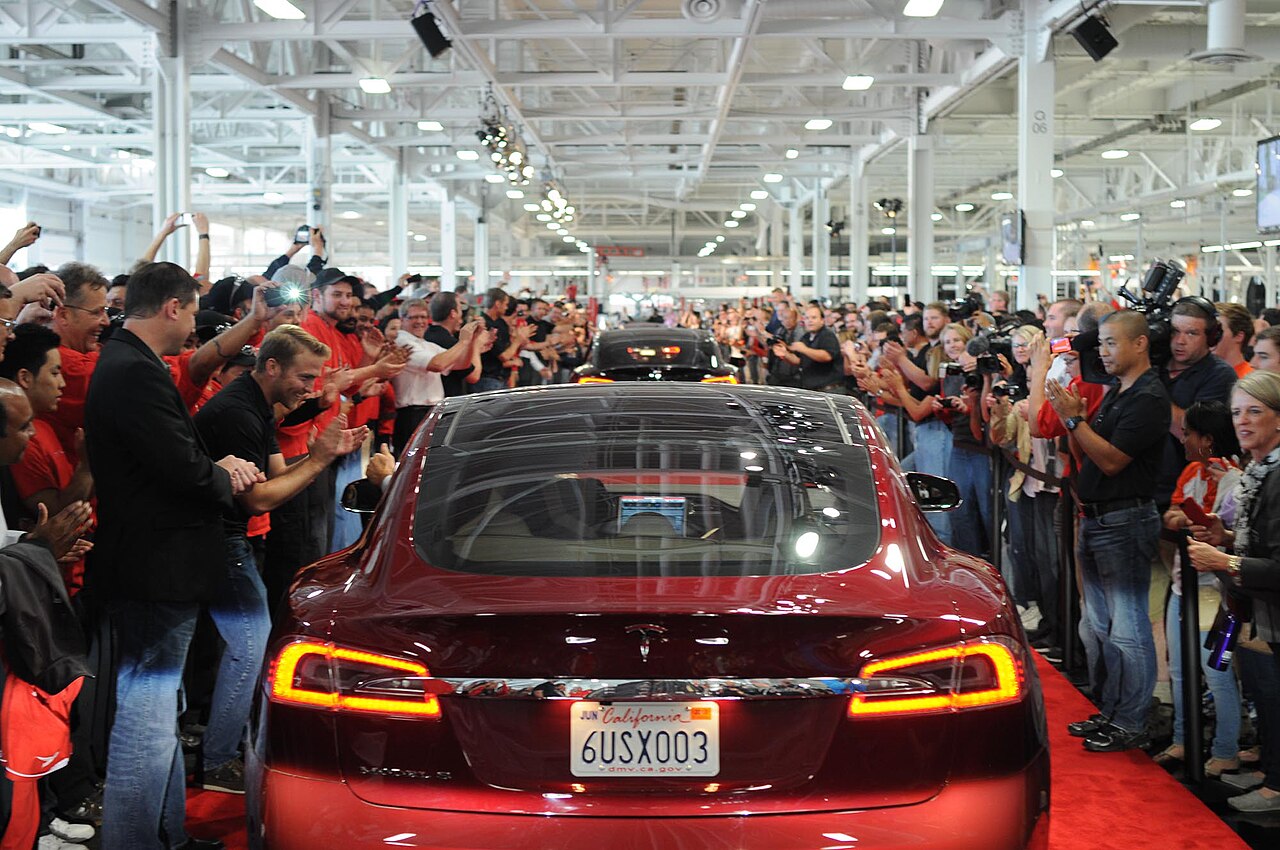 Um Tesla Model S vermelho, visto de trás, avança por um tapete vermelho dentro de uma fábrica, entre pessoas que aplaudem e fotografam.