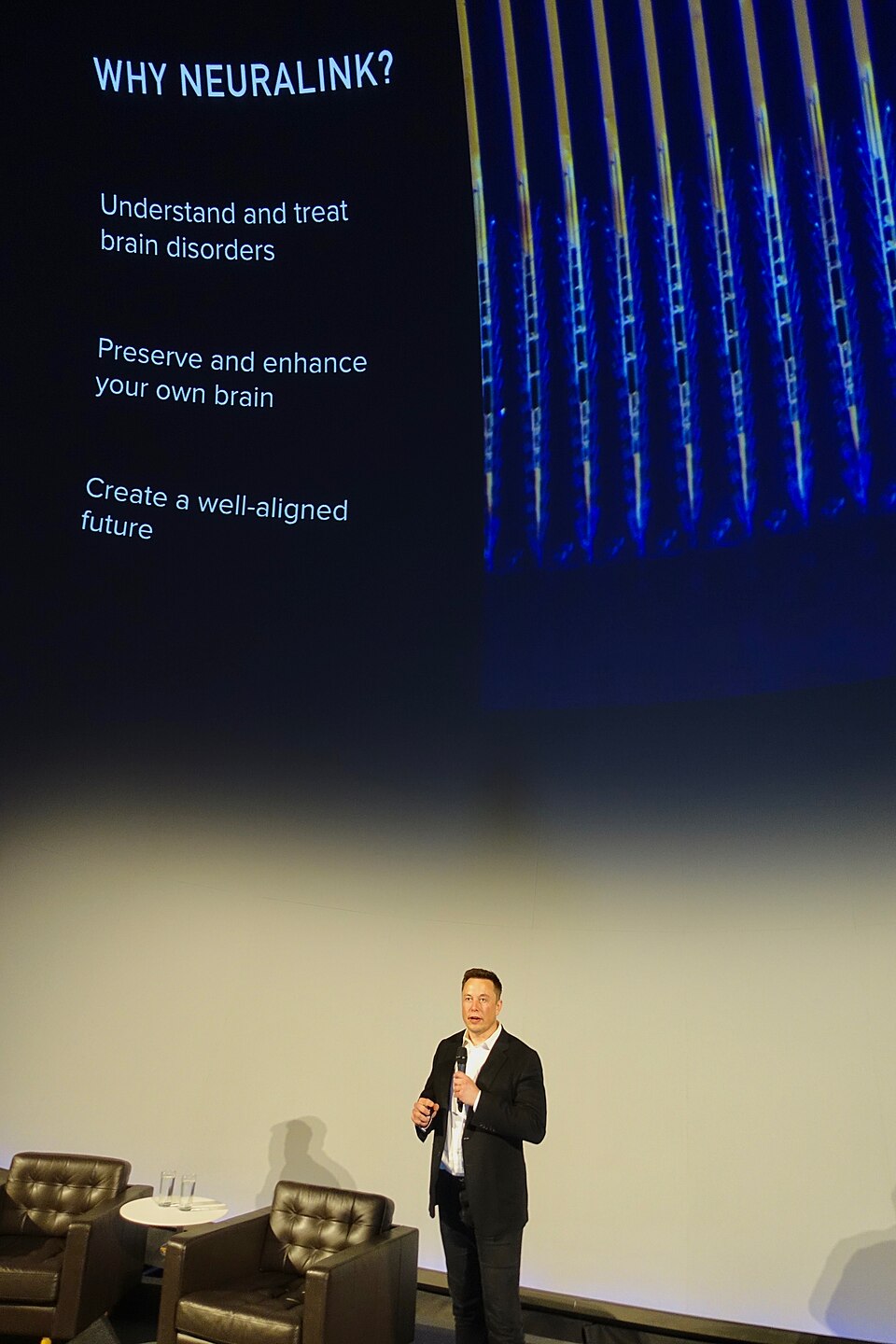 Elon Musk, de terno escuro, em pé num palco e segurando um microfone, diante de um telão com o título "Why Neuralink?" e uma lista de objetivos.