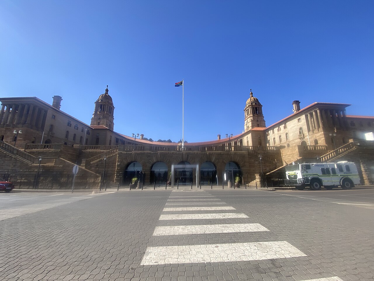 Fachada curva em arenito dos Union Buildings, com duas torres e uma bandeira ao centro, sob céu azul, vista de uma rua de paralelepípedos.