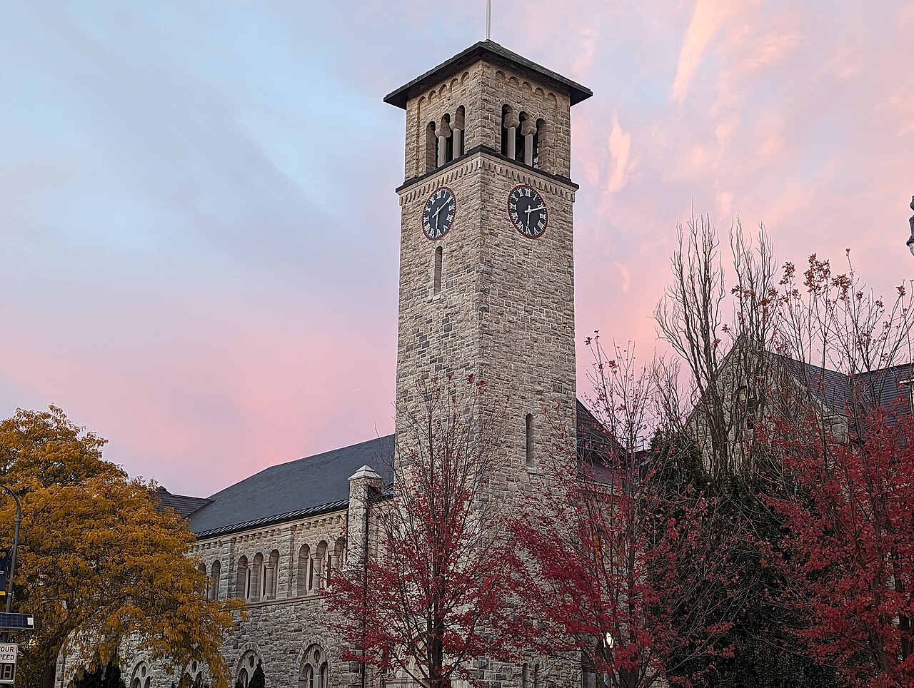 Torre de pedra com dois relógios do Grant Hall, no campus da Queen's University, entre árvores de outono, ao entardecer com céu rosado.