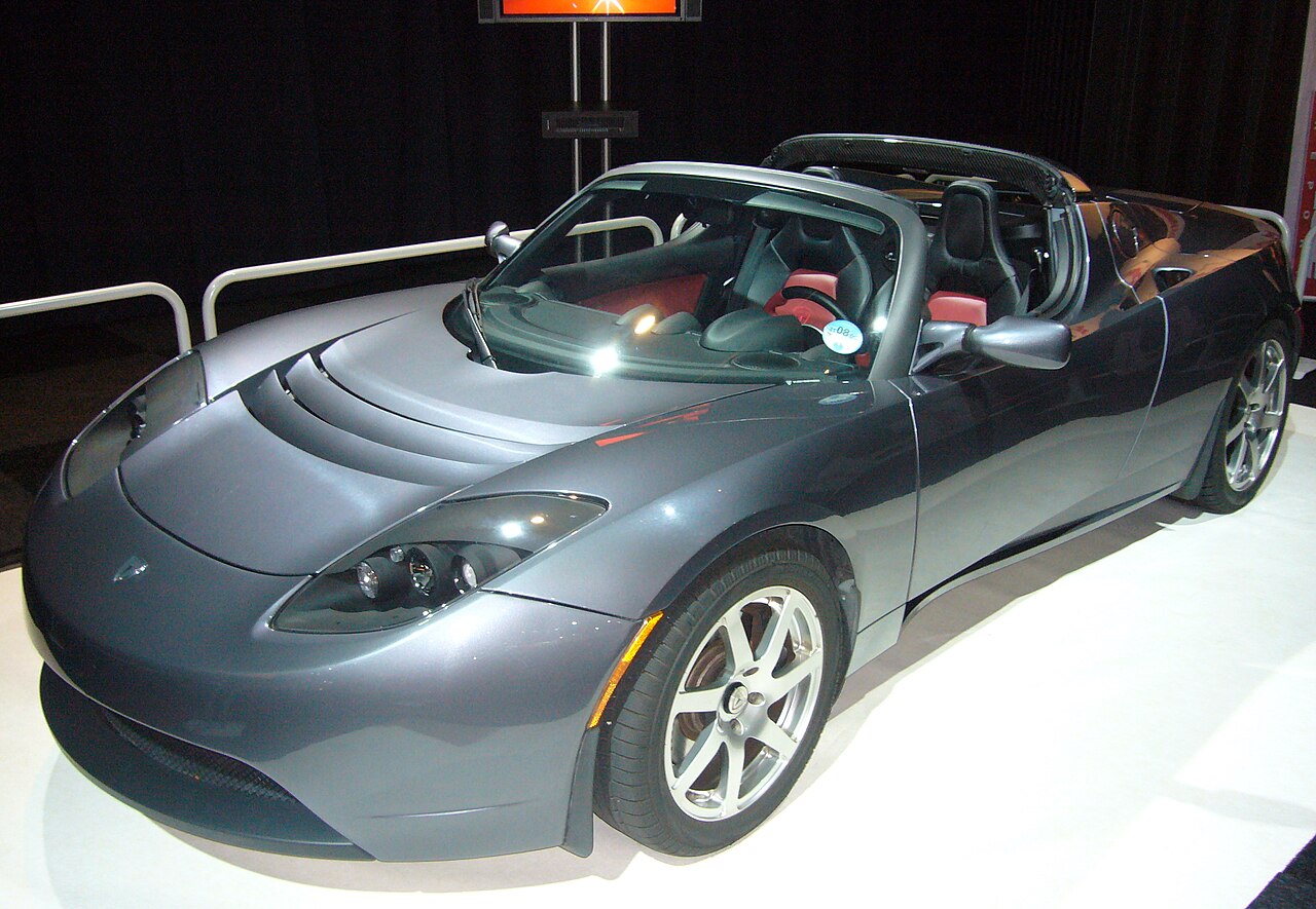 Esportivo Tesla Roadster cinza, conversível, com bancos vermelhos, exposto sobre um piso branco diante de uma cortina escura.