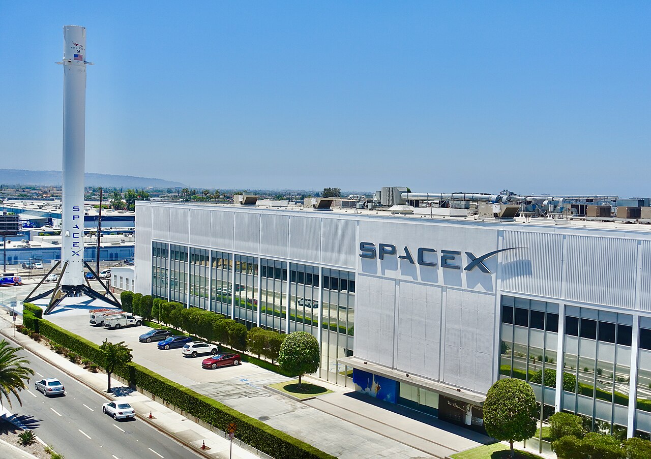 Prédio de fachada clara com o logotipo SpaceX e, ao lado, um primeiro estágio de foguete Falcon 9 em pé sobre suportes, sob céu azul.