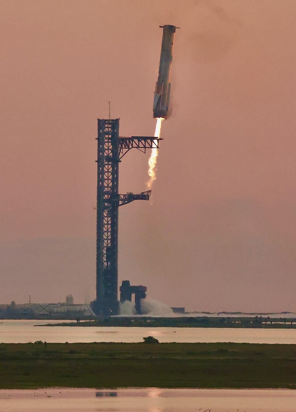 O propulsor Super Heavy, com a chama do motor acesa, descendo ao lado de uma alta torre de lançamento metálica, sob um céu rosado.