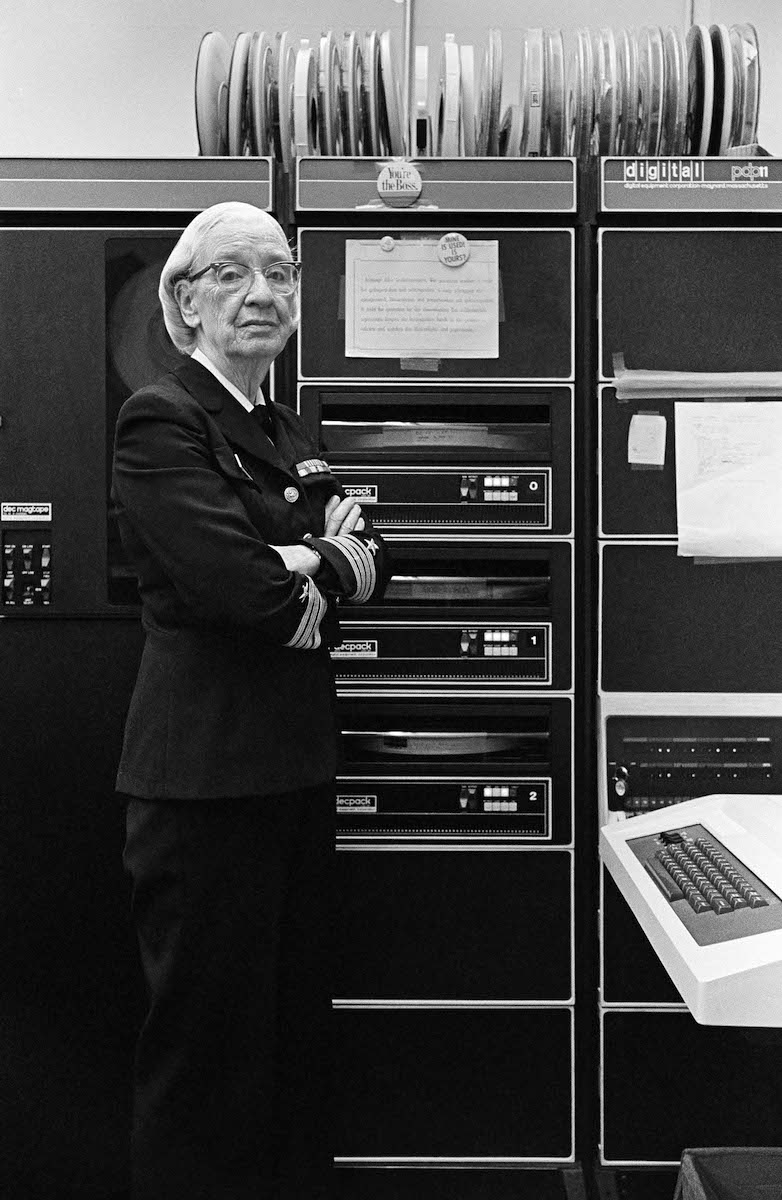Grace Hopper de uniforme da Marinha, em pé de braços cruzados ao lado de armários de um computador DEC PDP-11 e de unidades de fita magnética