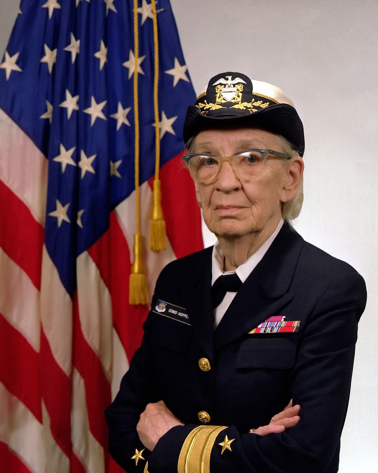 Grace Hopper de uniforme azul-marinho de comodoro, com quepe e óculos, braços cruzados, diante de uma bandeira dos Estados Unidos