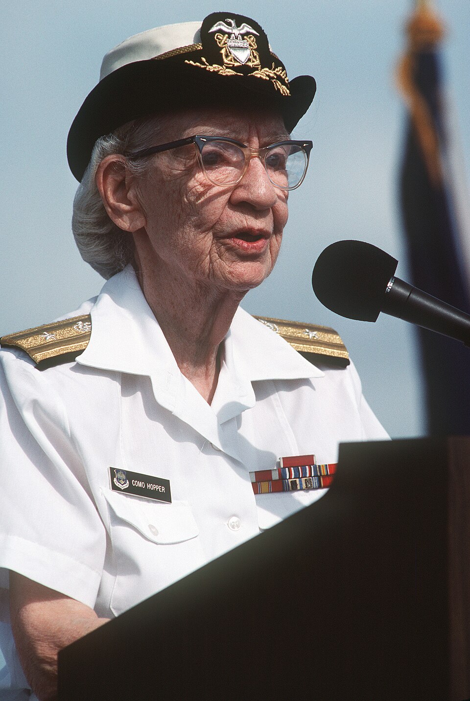 Grace Hopper de uniforme branco de verão e quepe, falando ao microfone num púlpito, com uma bandeira desfocada ao fundo