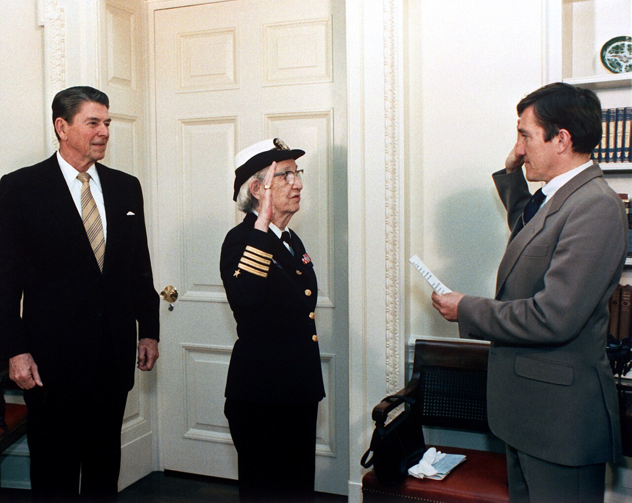 Grace Hopper de uniforme, com a mão direita levantada no juramento, entre o presidente Ronald Reagan, à esquerda, e o secretário da Marinha John Lehman, à direita
