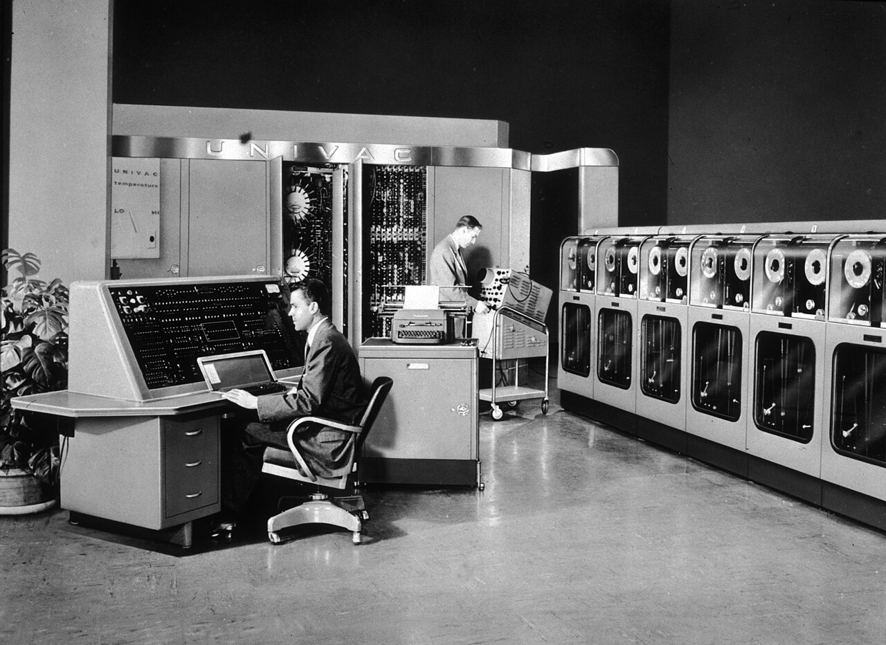 Dois homens operam o UNIVAC I: um sentado ao console de controle, outro em pé junto da unidade central, com uma fileira de unidades de fita magnética à direita