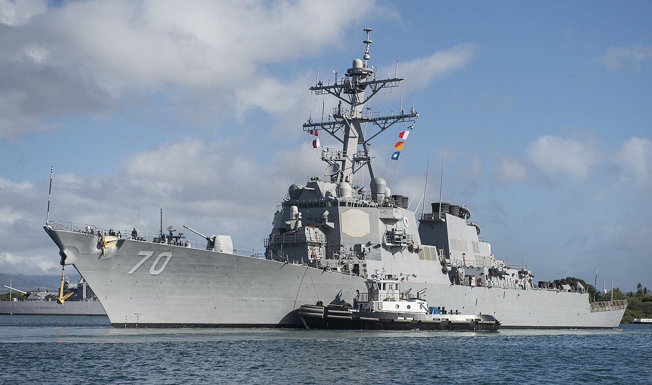 O destróier USS Hopper, casco cinza com o número 70 na proa, saindo do porto de Pearl Harbor com um rebocador ao lado e céu azul com nuvens