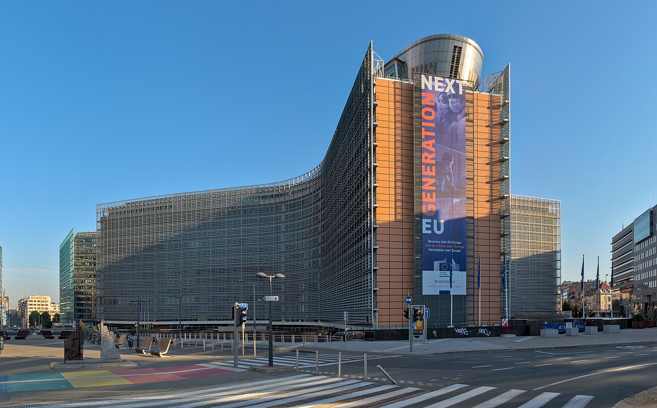 Edifício Berlaymont, sede da Comissão Europeia em Bruxelas, de fachada curva de vidro e metal, com uma faixa vertical azul e laranja, visto de uma esquina.
