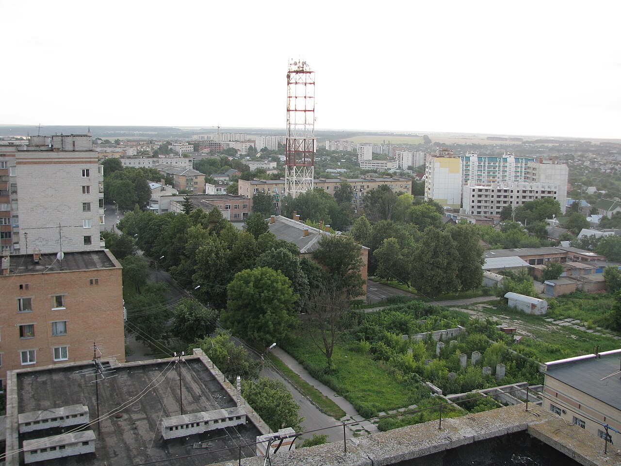 Vista de uma cidade ucraniana a partir de um prédio alto: blocos de apartamentos, árvores, uma torre de telecomunicações vermelha e branca e campos ao fundo.