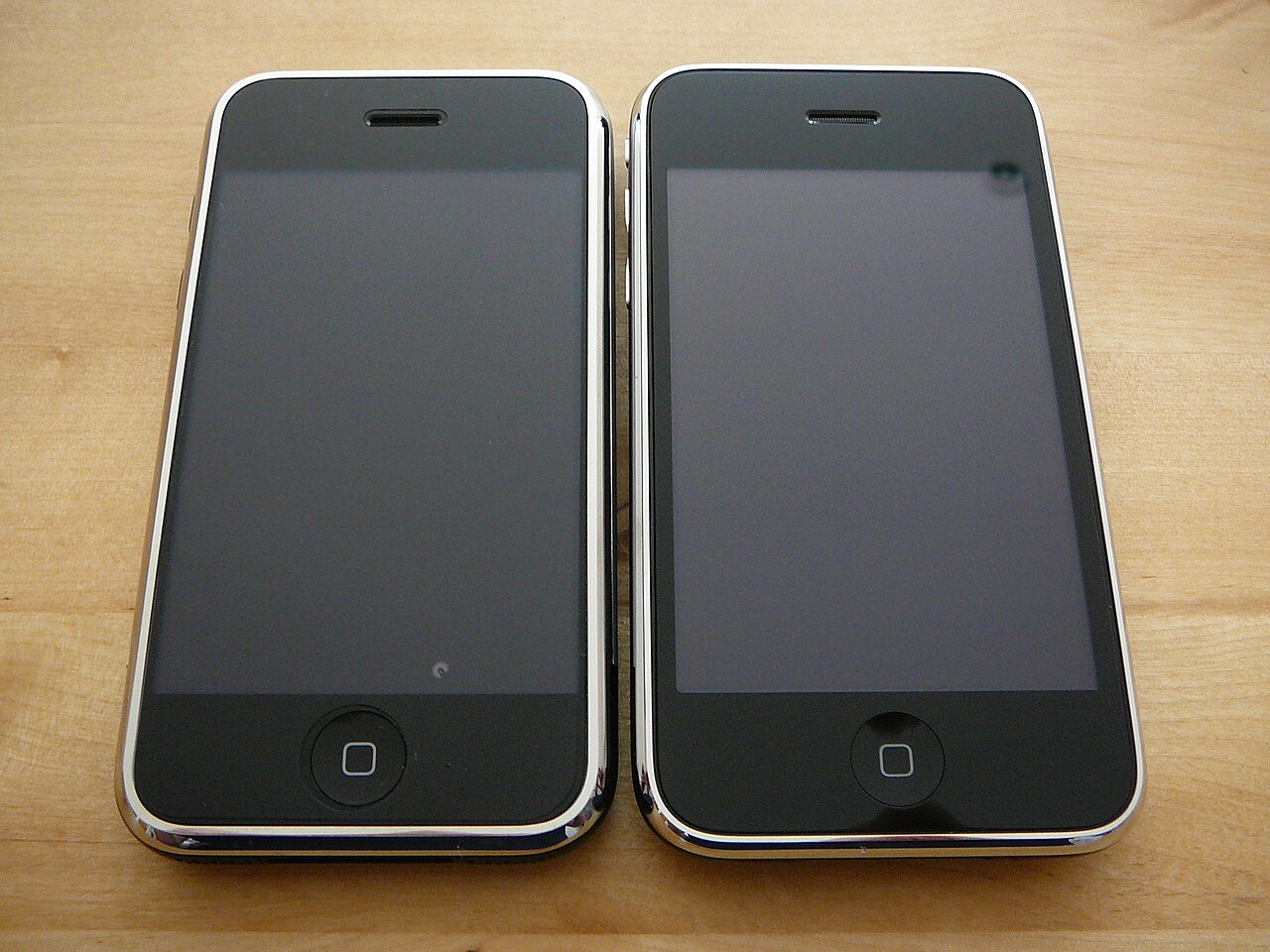 Dois iPhones lado a lado sobre uma mesa de madeira, vistos de cima e com as telas desligadas: o modelo original à esquerda e o iPhone 3G à direita.