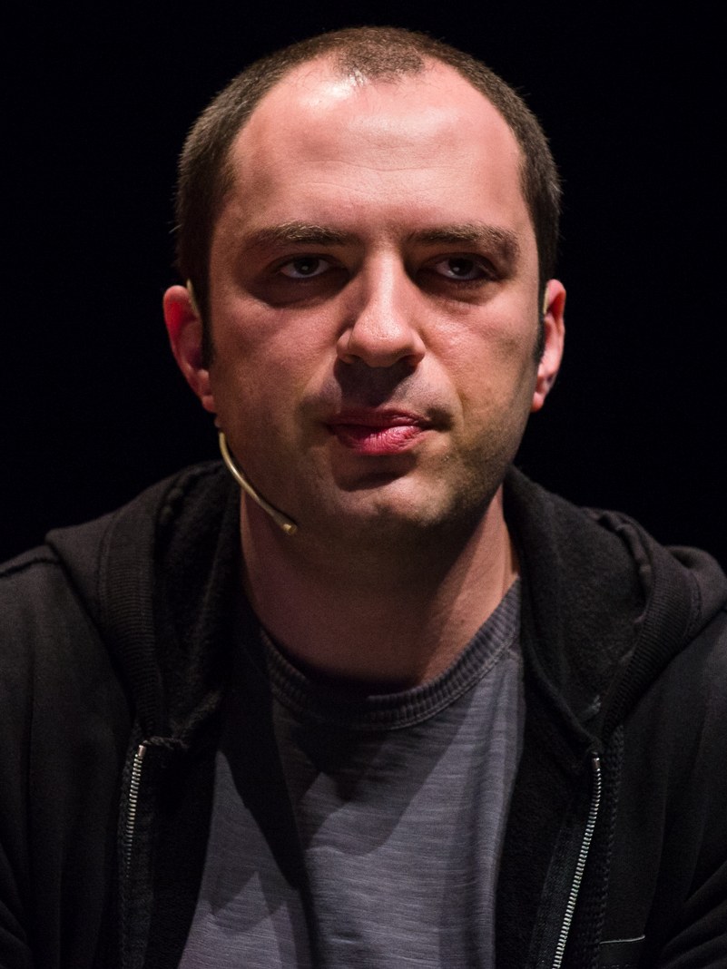 Close do rosto de Jan Koum diante de um fundo preto, com um microfone de headset junto ao rosto e moletom escuro sobre camiseta cinza.
