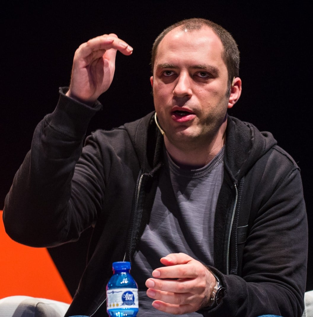 Jan Koum, de moletom preto e camiseta cinza, gesticula com a mão direita erguida diante de um fundo escuro, com um microfone de headset junto ao rosto.
