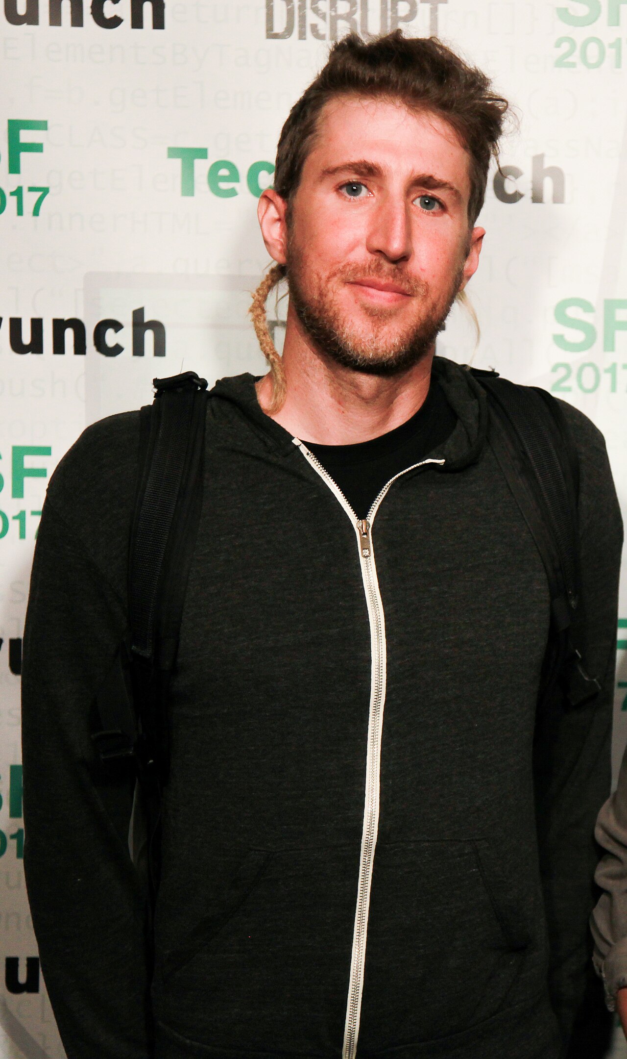 Moxie Marlinspike, de barba curta e cabelo preso, com moletom escuro de zíper aberto e mochila, diante de um painel de evento da TechCrunch.