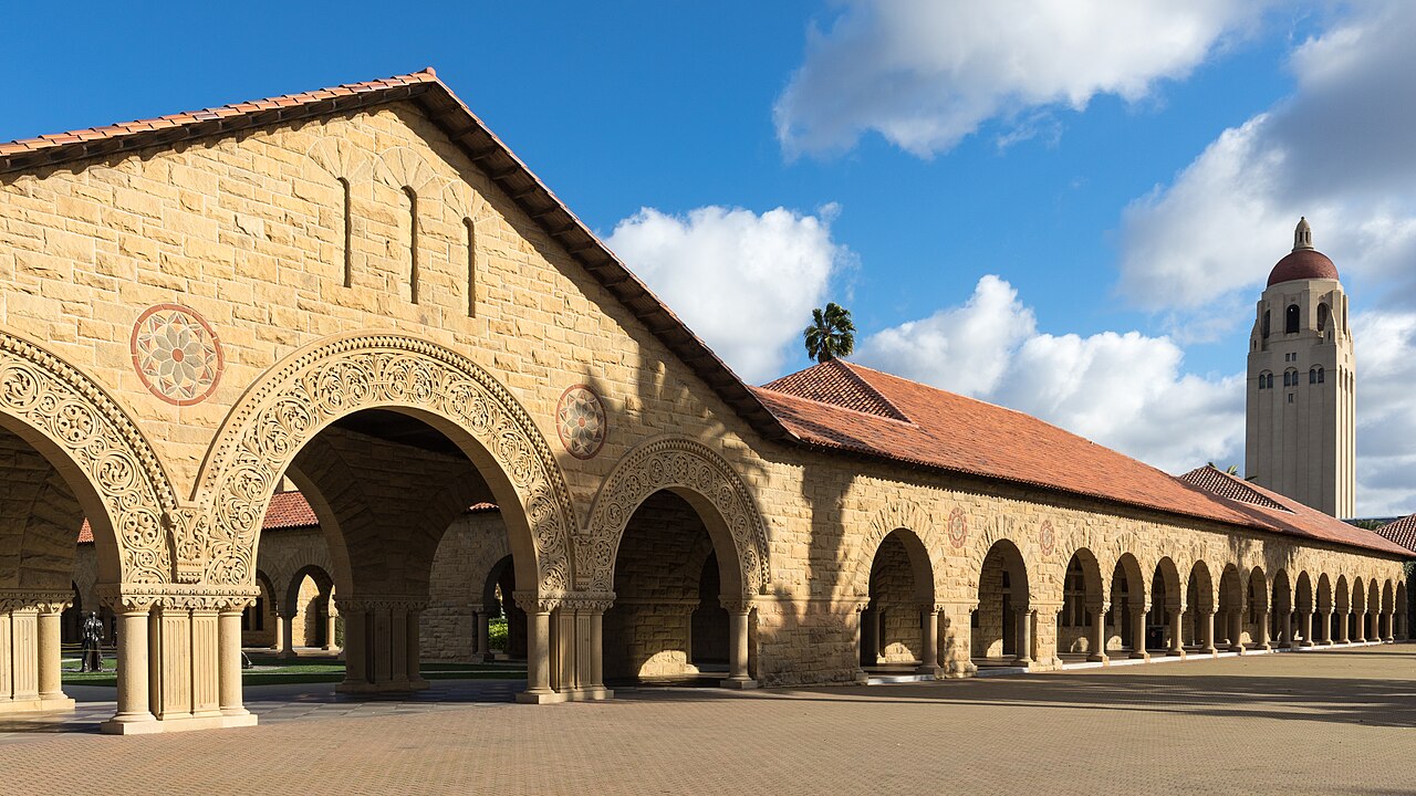 Arcadas de pedra clara com telhado de telhas vermelhas no campus de Stanford, com a torre Hoover à direita, sob céu azul com nuvens.