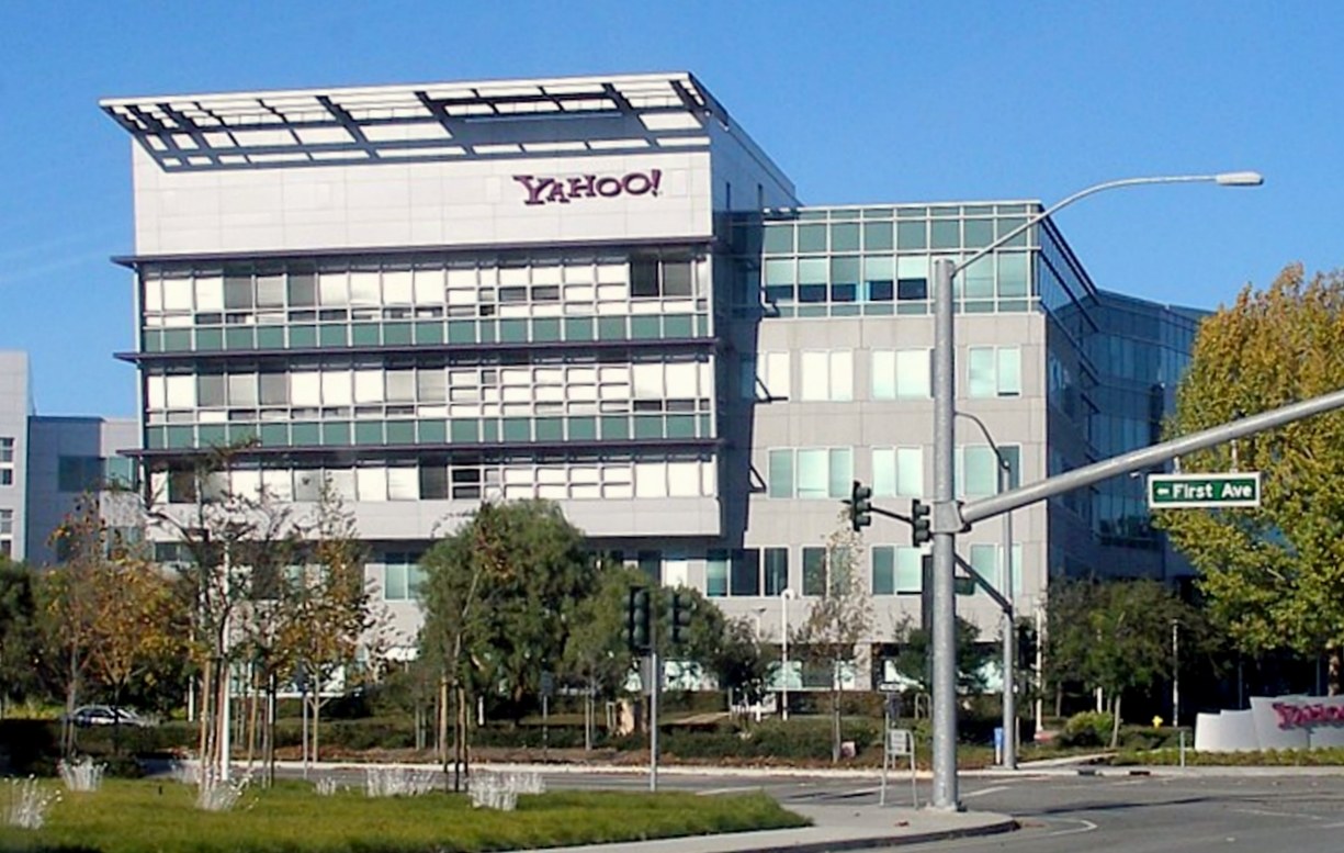 Prédio de escritórios de vários andares com fachada clara e janelas, o nome Yahoo em letras roxas no alto, sob céu azul, e um cruzamento de ruas à frente.