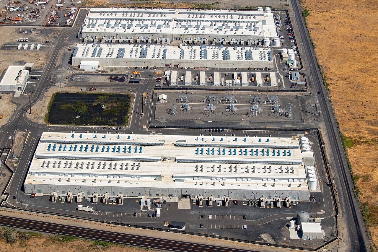 Vista aérea de três grandes prédios de data center de telhado plano e equipamentos de refrigeração, cercados por subestação elétrica e terreno seco.
